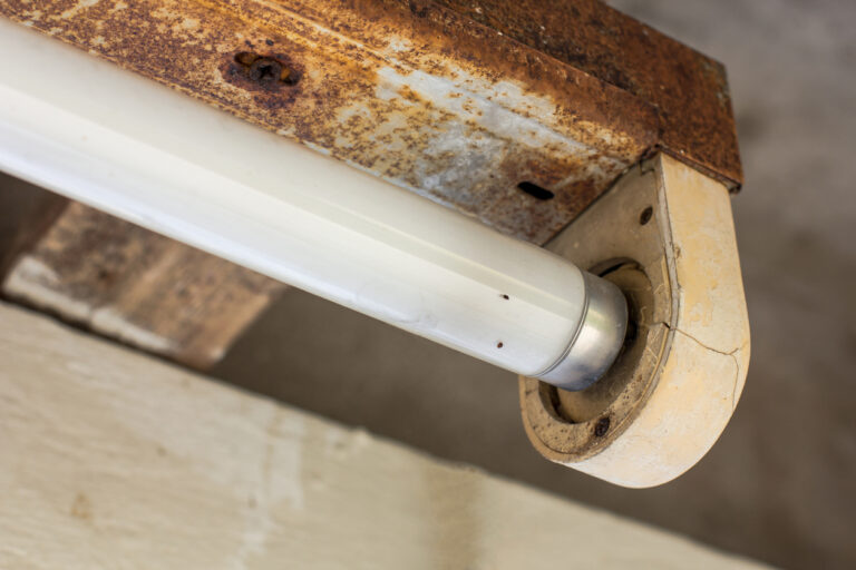 An electric lamp is placed in an open-air building. Rains, sunlight and heat erode the lamp rail but not the fluorescent bulb. This lamp is not in a good condition, but still functioning.