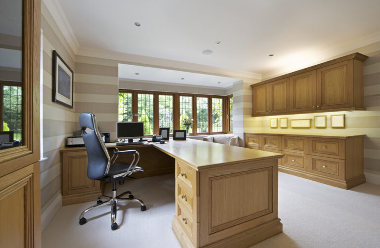 a large office in a luxury home with beautifully crafted light oak furnishings including desk and sideboard, cupboards, display cabinets and drawer chests. A large leather and chrome high-back chair sits to the left.