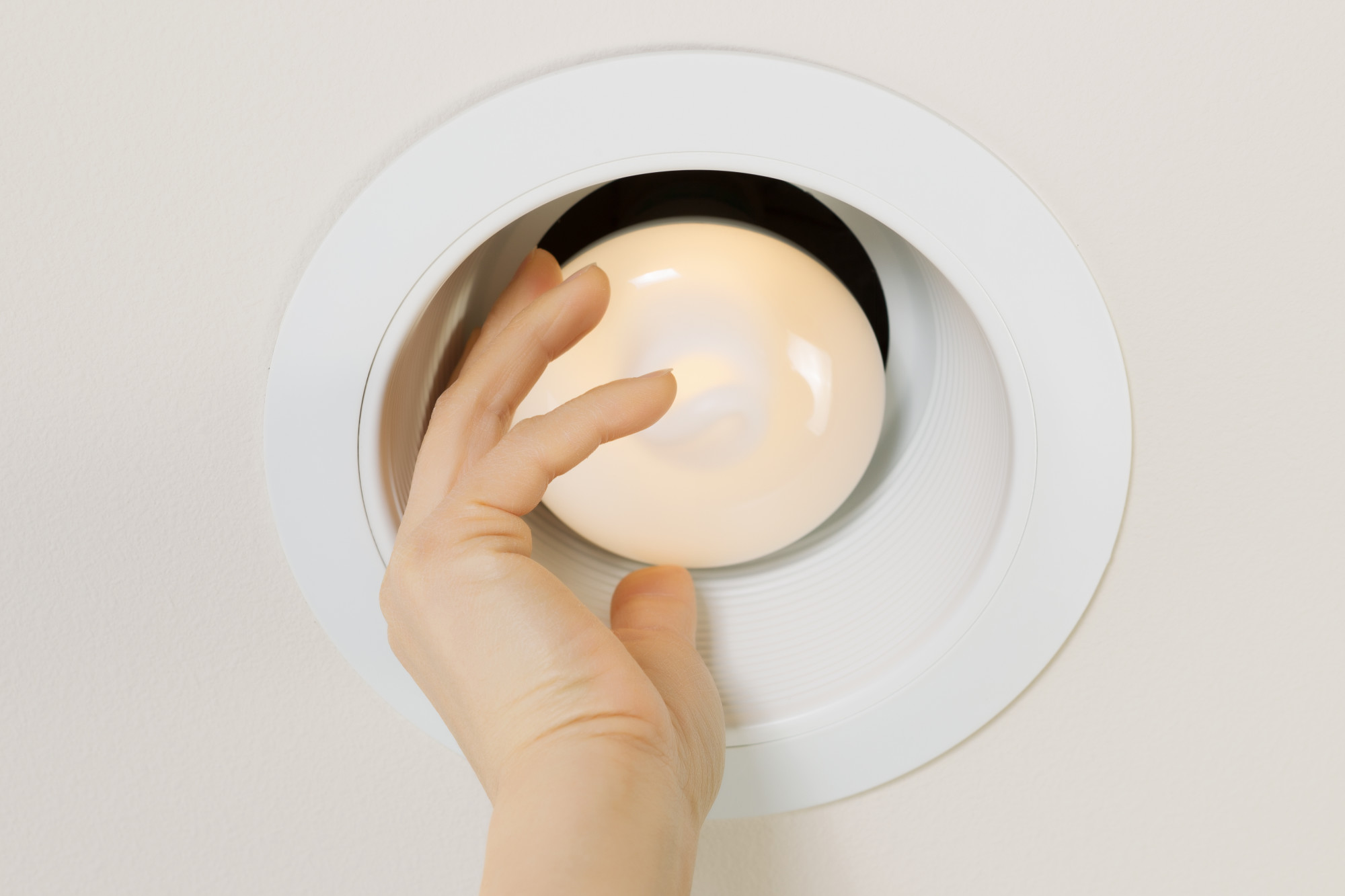 Photo of brand new illuminated flood light bulb being screwed in by female hand with recessed ceiling light mount in background